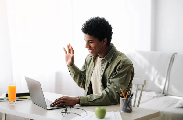 Distance communication concept. Positive black teen guy talking to his tutor or fellow students, waving at laptop webcam