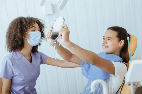 Menina pequena feliz olha no espelho para estimar o trabalho do dentista e desfrutar do resultado — Fotografia de Stock