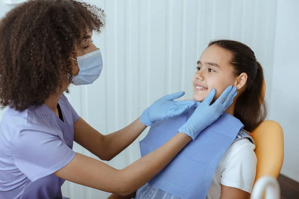 Giovane dentista afroamericano in uniforme, maschera protettiva e guanti esame dei pazienti bocca per prevenire la carie — Foto Stock