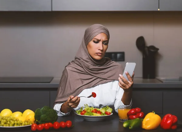 Szomorú muszlim nő telefonos étkezési saláta a modern konyha — Stock Fotó