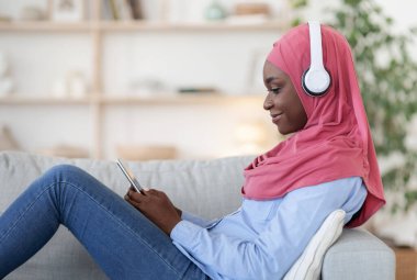 Kablosuz kulaklıklı siyah Müslüman kadın evde akıllı telefonuyla dinleniyor.