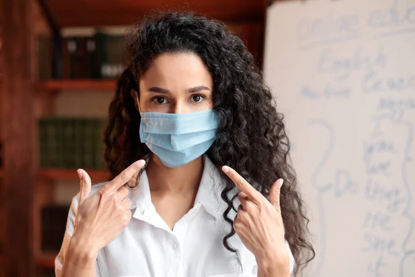 Учительница в медицинской маске и указывает на лицо — стоковое фото