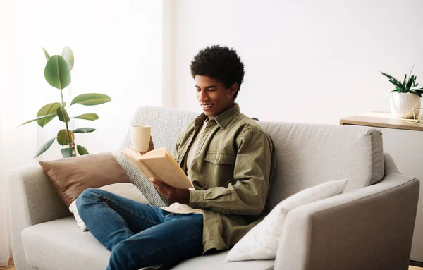 Vidám afro-amerikai tinédzser csésze forró kávé olvasás könyv kanapén otthon — Stock Fotó