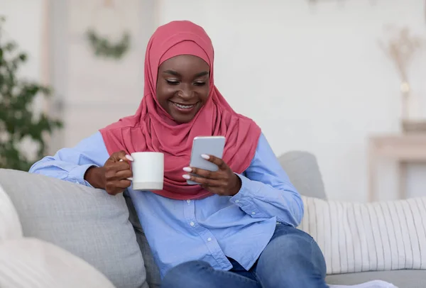 Fekete iszlám nő pihentető kanapén otthon Smartphone és kávé — Stock Fotó