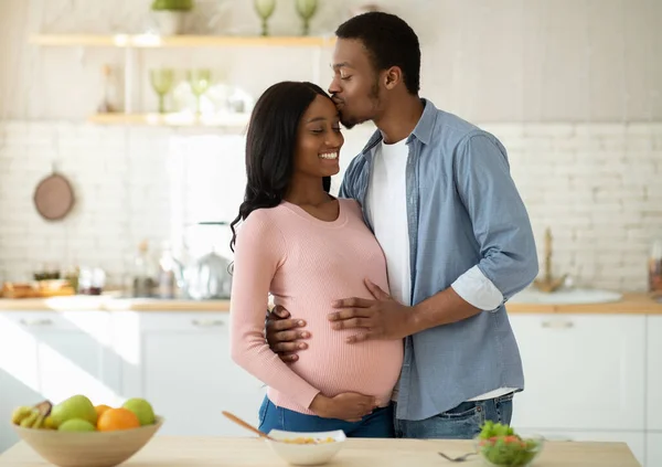Maternidad y concepto de alimentación saludable. Cuidar marido negro besando a su esposa embarazada en la cocina mientras se cocina el desayuno —  Fotos de Stock