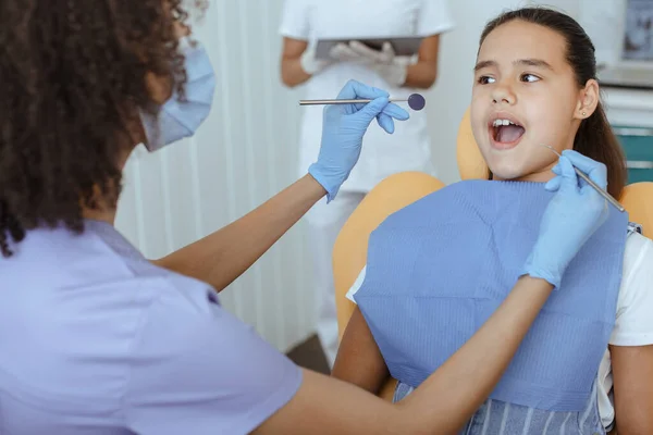 Young african american doctor in uniform, protective mask and gloves treats teeth of hispanic girl — Stock Photo, Image