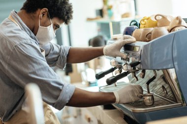 Bin yıllık Afro-Amerikan önlüklü, eldivenli ve koruyucu maskeli adam sabahları kahve makinesiyle iç mekanda çalışıyor.