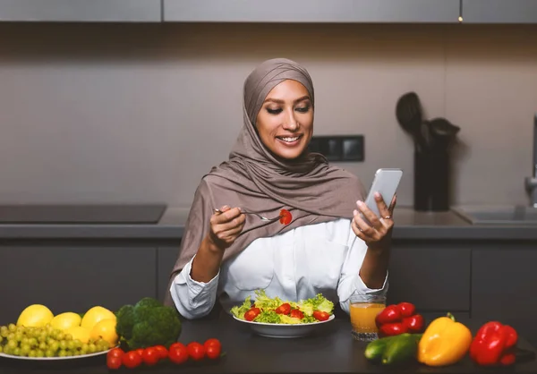 Muszlim nő használ okostelefon Evés Saláta Ebédelés a konyhában — Stock Fotó