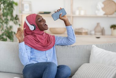 Siyahi Müslüman kadın evde eğleniyor, akıllı telefonda mikrofon gibi şarkı söylüyor.