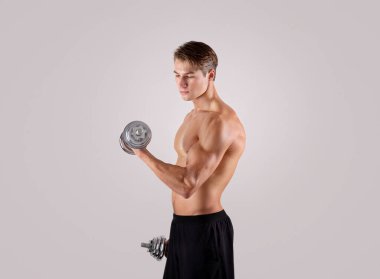 Yakışıklı beyaz vücut geliştirici, ağırlık pompalıyor, hafif stüdyo arka planında dambıllarla egzersiz yapıyor.