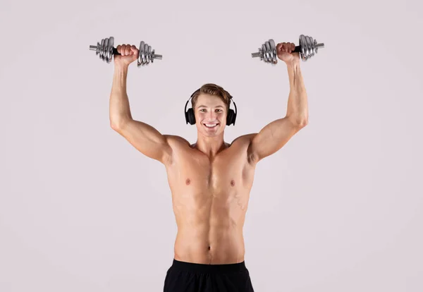 Jeune homme sportif avec torse nu soulevant des haltères et écoutant de la musique dans des écouteurs sur fond de studio léger — Photo