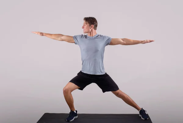 Schöner junger Mann in Sportbekleidung beim Krieger-Yoga-Pose auf Sportmatte vor hellem Hintergrund, Ganzkörperporträt — Stockfoto