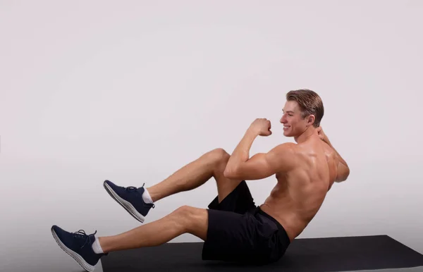 Feliz chico joven con el cuerpo muscular haciendo ejercicio en la alfombra deportiva sobre fondo de estudio de luz —  Fotos de Stock