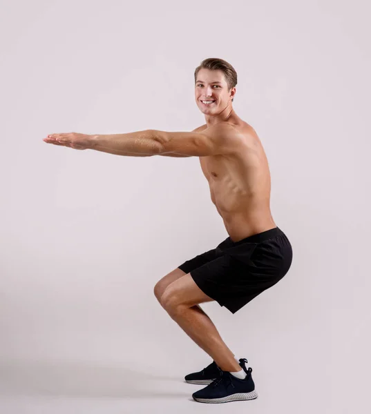 Retrato de cuerpo entero de un chico sexy sin camisa haciendo sentadillas, mirando a la cámara y sonriendo en el fondo del estudio de luz — Foto de Stock