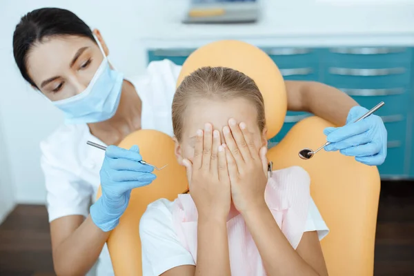 Little child covers face with hands, sits in medical chair with woman doctor holds instruments for examining patient — Stock Photo, Image