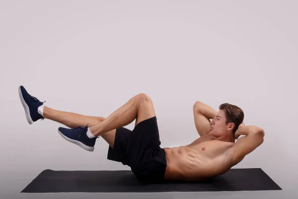 Starker junger Kerl mit nacktem Oberkörper macht Bauchmuskelübungen auf Yogamatte vor hellem Studiohintergrund, Seitenansicht — Stockfoto