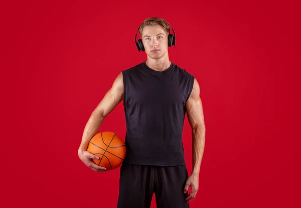 Jogador de basquete millennial atraente ouvir música, usando fones de ouvido sobre fundo estúdio vermelho — Fotografia de Stock