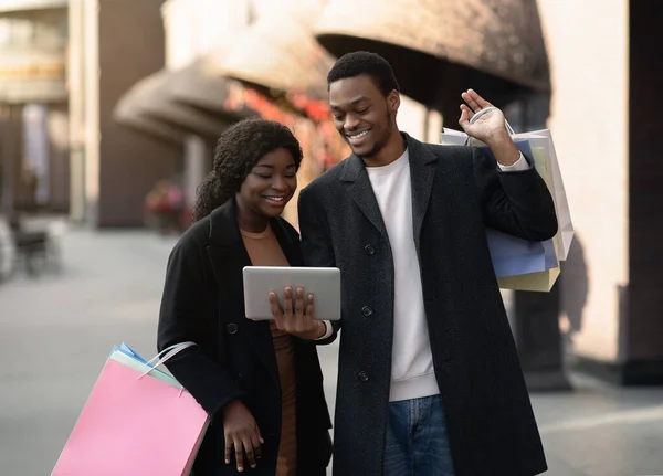 Enthousiaste homme afro-américain millénaire en manteaux détient des sacs à provisions et montre tablette numérique à sa femme — Photo