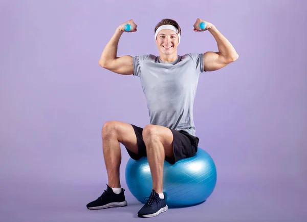 Glad ung idrottsman sitter på fitness boll och träna med hantlar på violett studio bakgrund, full längd — Stockfoto