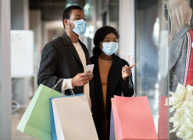 Mutlu, Afro-Amerikalı, koruyucu maskeli bir çift alışveriş merkezinde yürüyor, renkli çantalar taşıyor.