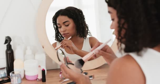 Trucco mattutino. Giovane donna afro-americana che applica il rossetto con la spazzola sulla guancia, seduta davanti allo specchio a casa — Video Stock