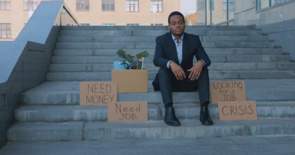 Deprimerad afrikansk amerikansk man sitter i förtvivlan på trappor med låda med personal och plakat Behöver jobb — Stockvideo