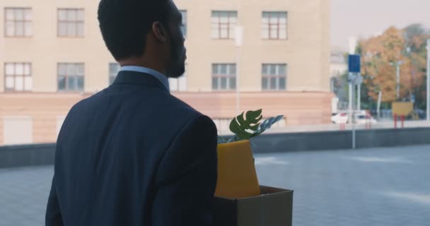 Уволен африканский офисный работник, выходящий на улицу с коробкой личных вещей, расстроен после увольнения — стоковое видео