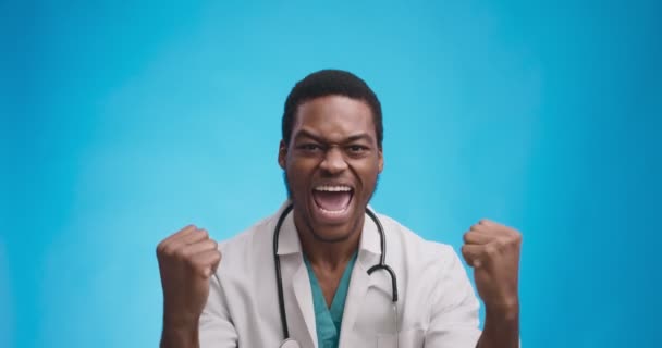 Feliz emocionado médico afroamericano animando y celebrando el éxito y ganar, fondo de estudio azul — Vídeos de Stock