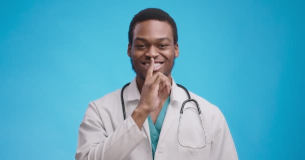 खुश अफ्रीकी अमेरिकी चिकित्सा डॉक्टर चुप्पी इशारा दिखा रहा है, होंठों पर उंगली डाल रहा है और कैमरा को मुस्कुरा रहा है — स्टॉक वीडियो