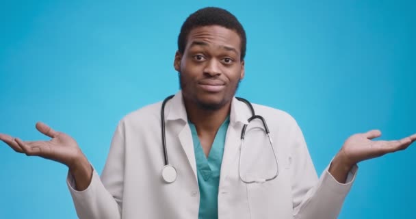 Retrato de médico afro-americano inseguro em brasão médico branco encolhendo os braços, fazendo eu não sei gesto — Vídeo de Stock