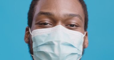 Koruyucu tıbbi maske takan, gülümseyen ve kafa sallayan, mavi stüdyo geçmişi olan Afro-Amerikan bir adamın yakın portresi.