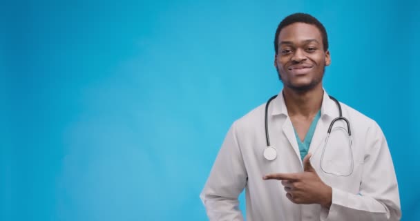 खुश अफ्रीकी अमेरिकी डॉक्टर कॉपी स्पेस पर एक तरफ इशारा करते हुए और मुस्कुराते हुए, नीले स्टूडियो पृष्ठभूमि — स्टॉक वीडियो