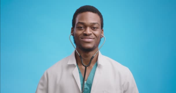 Retrato de un joven médico sonriente en una bata blanca médica profesional que señala el estetoscopio a la cámara, fondo azul — Vídeos de Stock