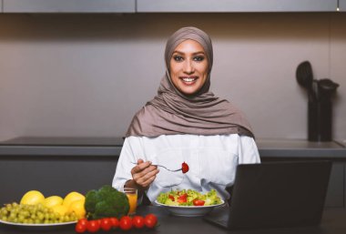 Arap kadın yemek yiyor Mutfaktaki dizüstü bilgisayarın başında oturuyor