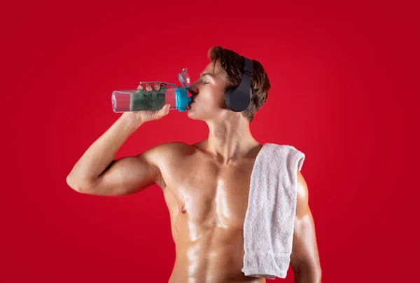 Sexy joven deportista con torso desnudo y toalla escuchando música en eadphones y agua potable sobre fondo rojo — Foto de Stock