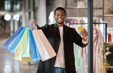 Binlerce yıllık neşeli Afro-Amerikan adam kredi kartıyla poz veriyor ve alışveriş merkezinde renkli çantalar alıyor.