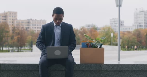 Fired black entrepreneur in suit using laptop for online job searching, sitting with box of personal belongings outdoors — Stock Video