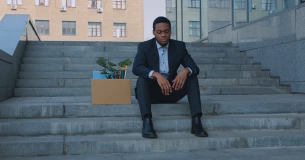 Отчаянный африканский американский служащий, сидящий на лестнице с коробкой личного персонала после увольнения, отслеживающий выстрел — стоковое видео