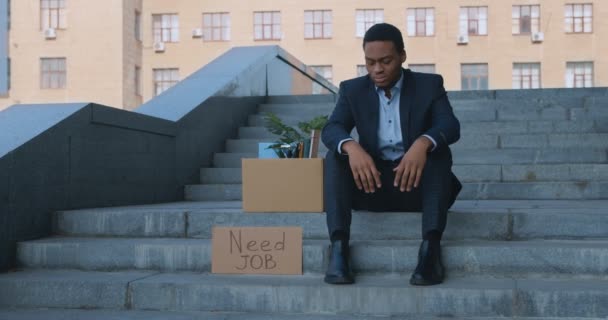 Отчаянный африканский американский бизнесмен, сидящий рядом с офисным центром с плакатом "Нужна работа, провал бизнеса" — стоковое видео