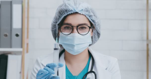 Primer plano retrato del médico en máscara médica y sombrero desechable empujando la jeringa con la vacuna, mirando a la cámara — Vídeos de Stock