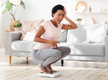 Aşırı kilolu olmaktan korkan duygusal Afrikalı Amerikalı kadın, şokla terazide oturuyor, kapalı alanda.