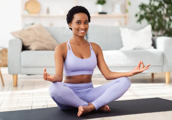 Улыбающаяся черная женщина в спортивной одежде сидит на коврике для йоги в позе лотоса и медитирует или делает дыхательные упражнения в помещении — стоковое фото