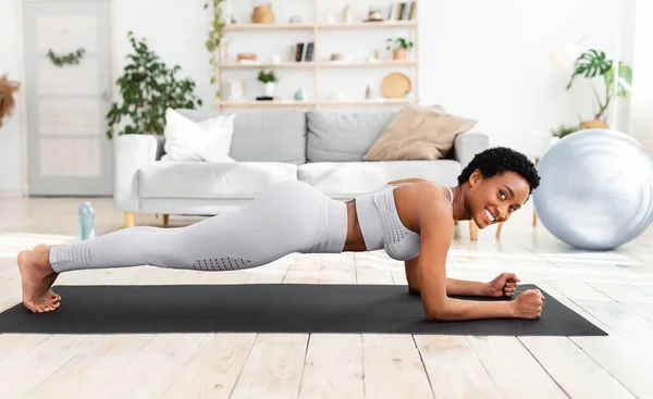 Hermosa mujer negra haciendo entrenamiento de fitness en alfombra deportiva en casa, de pie en la tabla del codo, el fortalecimiento de los músculos abdominales —  Fotos de Stock