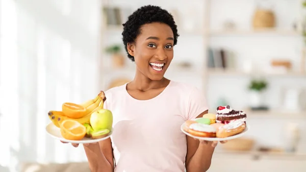 Felice signora nera in possesso di piatti con frutta e dolci a casa, facendo la scelta tra cibi sani e malsani — Foto Stock