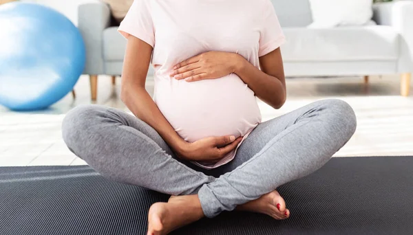 認識できない黒妊娠中の女性座っています蓮のポーズでヨガマットと巨大彼女の大きなおなかで家,パノラマ — ストック写真