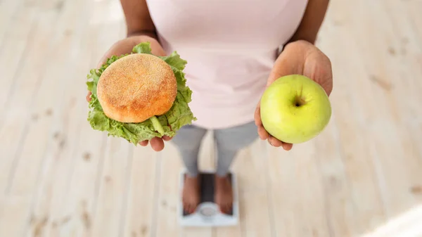 Вид зверху чорношкіра жінка тримає яблуко і гамбургер, стоїть на масштабах, роблячи вибір своєї дієти, крупним планом — стокове фото