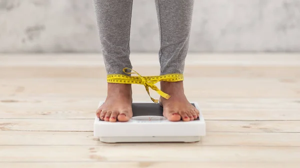 Unrecognizable black woman standing on scales, checking her weight with legs tied by tape measure
