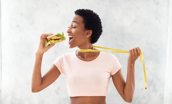 Desglose de dieta. Negro señora tratando de comer hamburguesa malsana, tirando de sí misma por cinta métrica, fondo gris —  Fotos de Stock