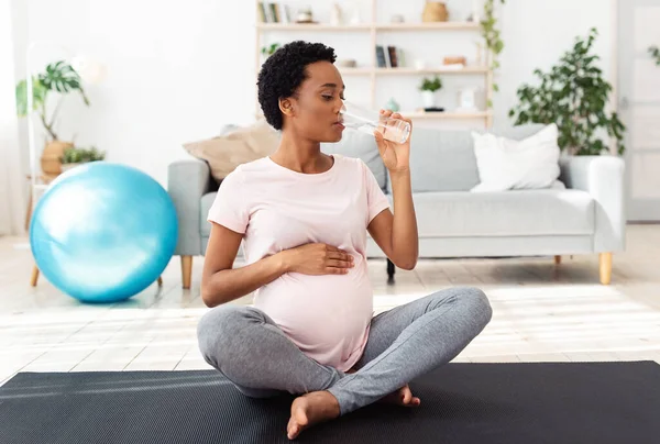 Nawadniaj się podczas ciąży. Czarna oczekująca pani pijąca szklankę wody podczas treningu na macie do jogi w domu — Zdjęcie stockowe