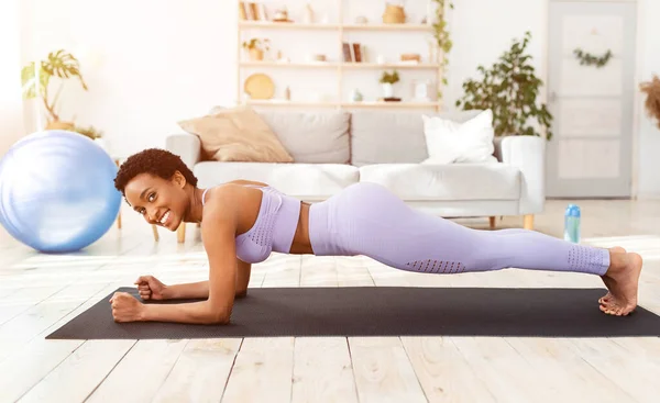 Deportes y pérdida de peso en casa. Atlética mujer afroamericana haciendo ejercicios sobre estera de yoga en el interior del salón — Foto de Stock